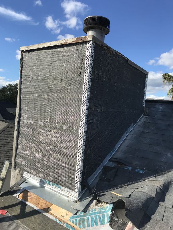 A chimney under construction or repair, covered in black mesh and metal corner bead by Stucco Services Central Florida, sits on a shingled roof with a clear blue sky and scattered clouds in the background.