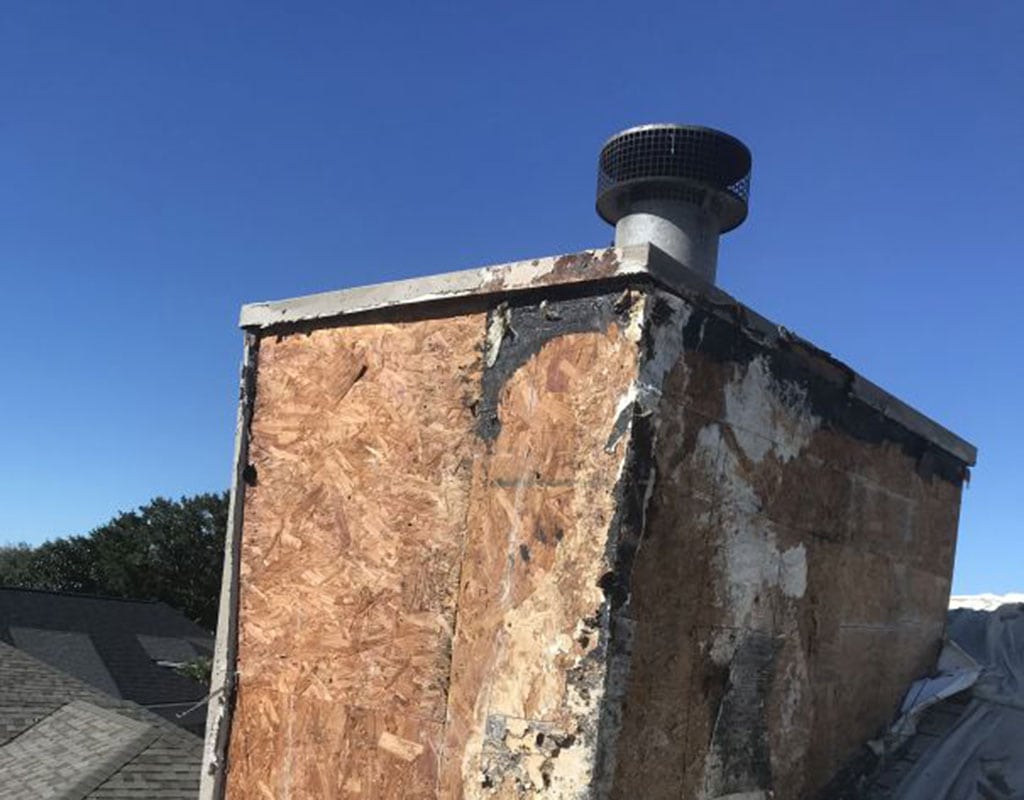 A weathered, partially damaged chimney with exposed wood and worn materials is shown on a rooftop under a clear blue sky—an ideal candidate for Stucco Services Central Florida.