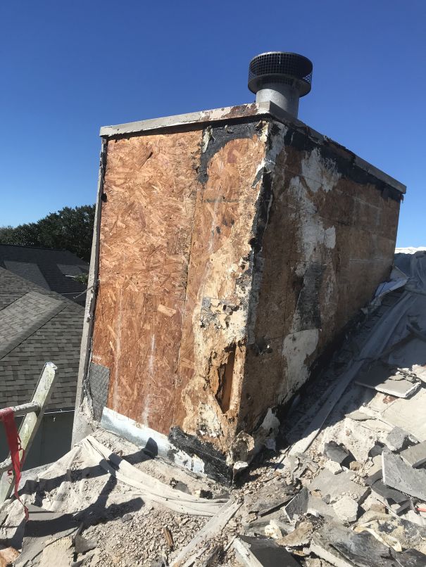 A heavily damaged chimney stands on a sloped roof, with missing and deteriorating exterior materials exposing plywood and crumbling sections—an issue often addressed by Stucco Services Central Florida. Debris and broken shingles scatter beneath a clear blue sky.