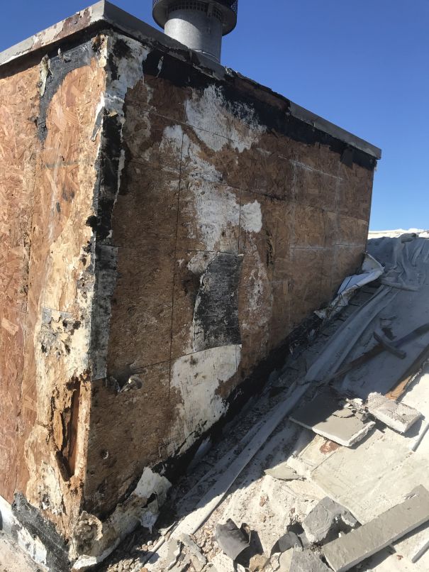 A damaged chimney with exposed plywood and peeling, blackened material stands on a rooftop under a clear blue sky. The surrounding roofing appears deteriorated and partially torn, highlighting the need for expert Stucco Services Central Florida.