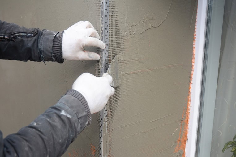 A person wearing white gloves applies plaster to a wall using a trowel, smoothing the surface around a metal corner bead near a window—a detail of expert work provided by Stucco Services Central Florida.