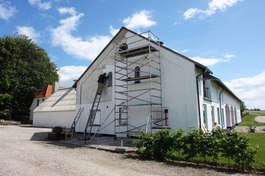 A white two-story house with scaffolding and a ladder set up on one side, where two workers from Stucco Services Central Florida are performing maintenance or repairs under a bright blue sky with scattered clouds.