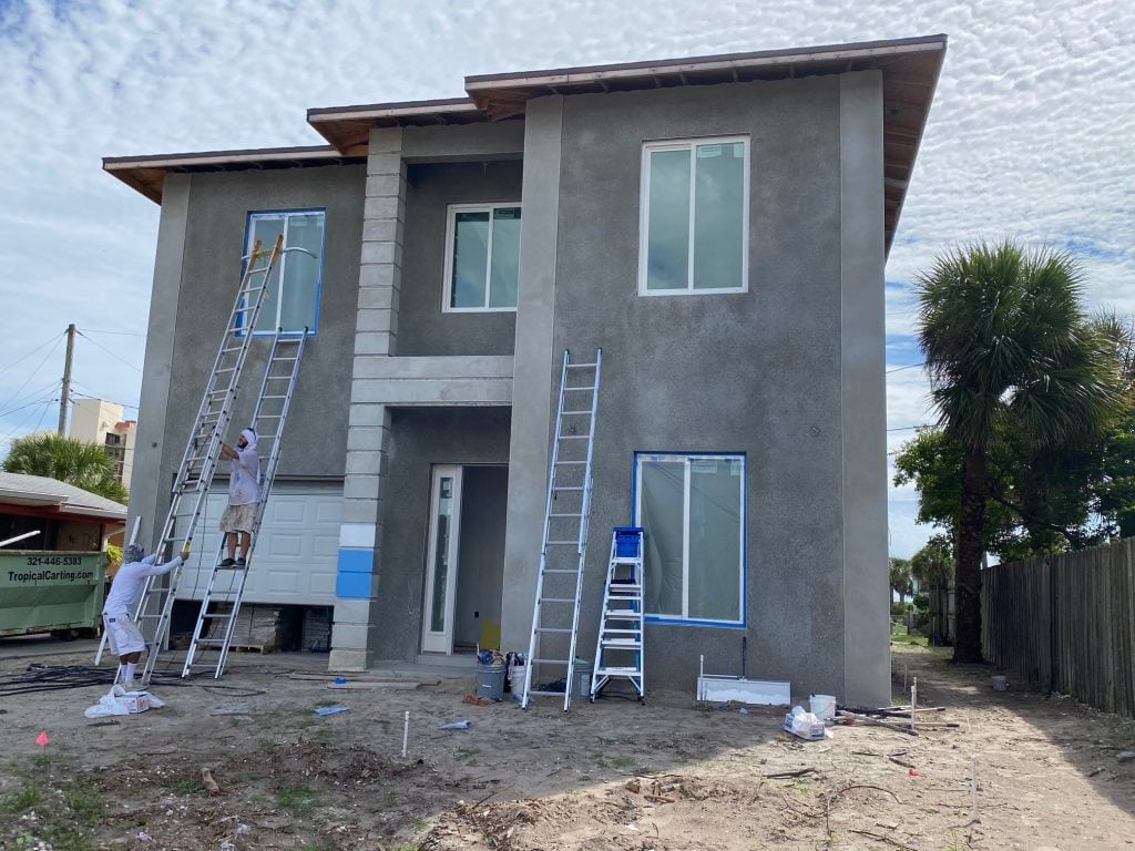 Two painters on ladders work on painting the exterior of a modern two-story house. The ground is bare, with scattered painting supplies, and the windows are taped for protection. A palm tree stands beside this home receiving Stucco Services Central Florida.