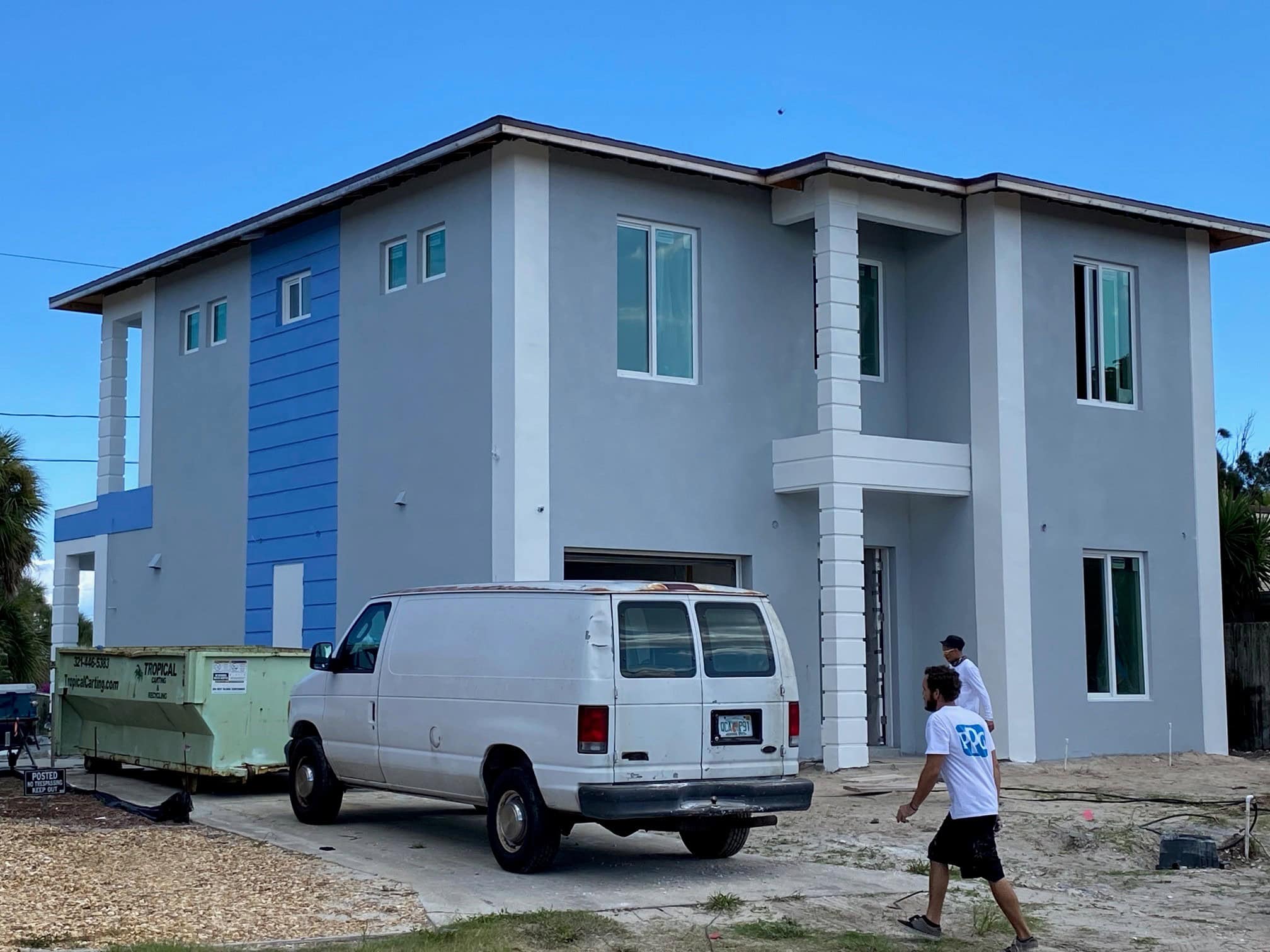 A modern two-story gray and blue house with large windows showcases fresh updates. A white van is parked in the driveway alongside a green dumpster, suggesting ongoing renovation or Stucco Services Central Florida involvement. Two people walk nearby.