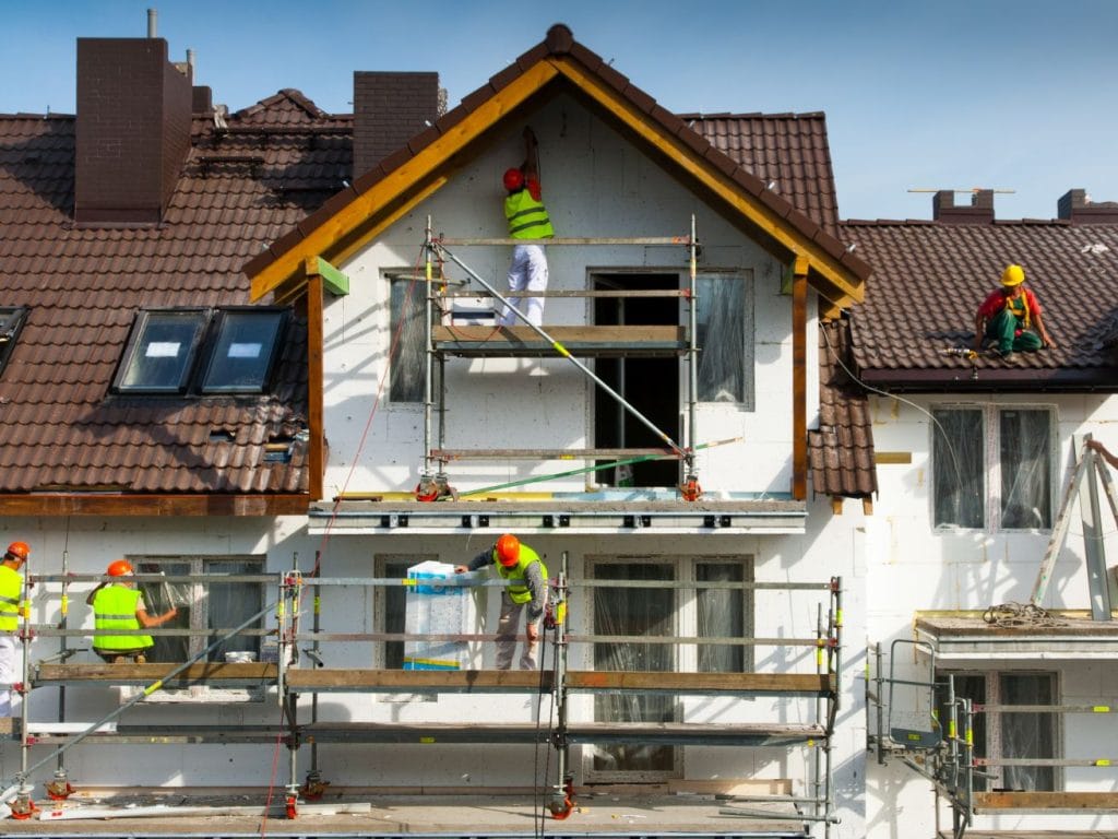 Construction workers in safety vests and helmets work on scaffolding, repairing and painting the exterior of a house with a brown-tiled roof—showcasing expert Stucco Services Central Florida—on a sunny day.