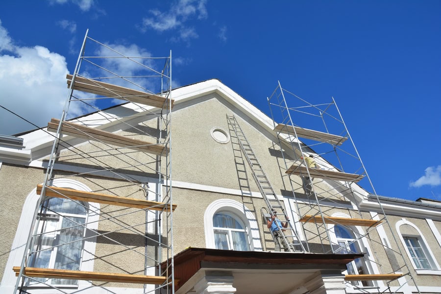 Scaffolding and a ladder are set up against the exterior of a light-colored building under a clear blue sky. A worker from Stucco Services Central Florida stands on the scaffolding, appearing to work on the building’s upper facade.