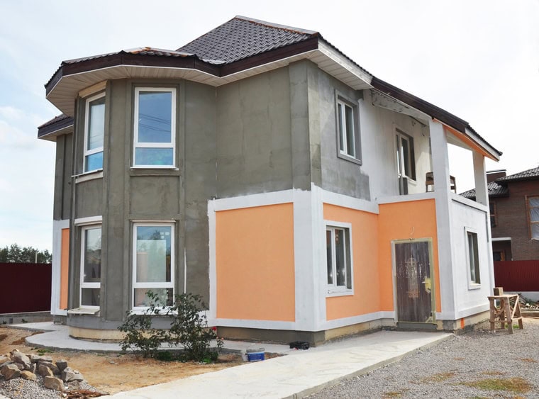 Two-story house under construction with gray upper walls, orange lower walls finished by Stucco Services Central Florida, white trim, bay windows, and a dark tiled roof. The yard has unfinished landscaping and construction materials nearby.