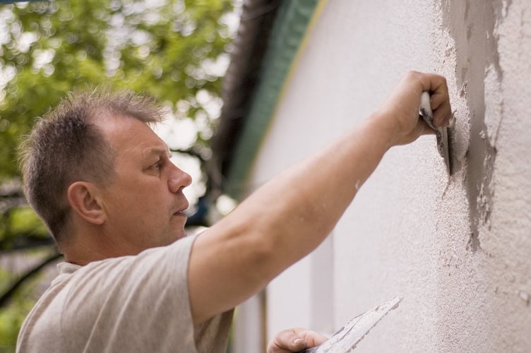A man in a light shirt applies plaster or cement to an exterior wall with a trowel, showcasing Stucco Services Central Florida expertise while working outdoors beside a building with trees in the background.