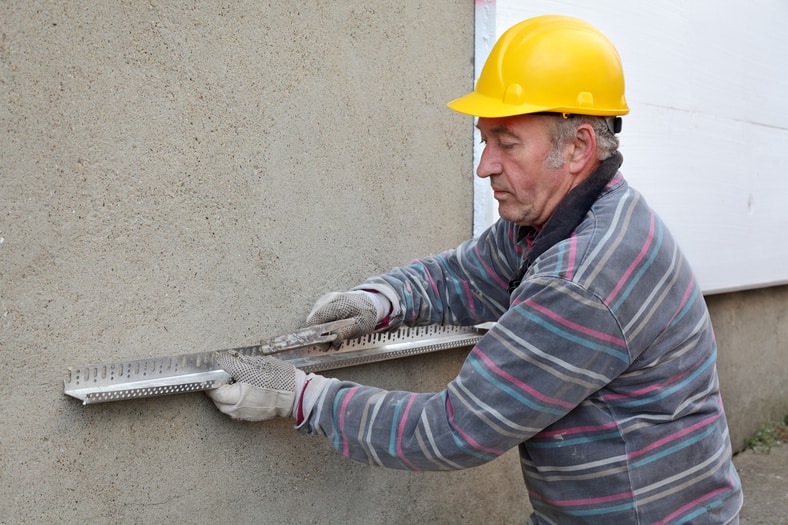 A construction worker wearing a yellow hard hat and gloves uses a tool to measure or align a metal strip on a textured exterior wall, showcasing expert work typical of Stucco Services Central Florida.