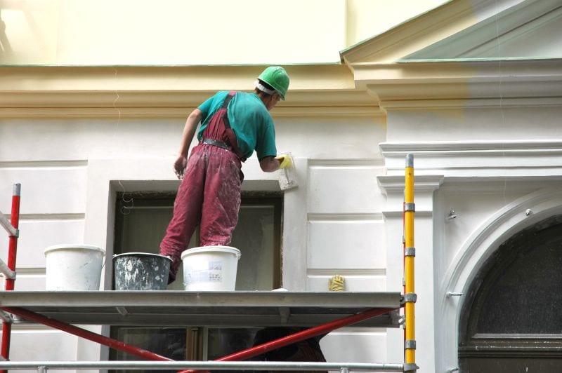 A construction worker wearing a green helmet and red overalls stands on scaffolding, painting the exterior wall of a building with a brush—showcasing expert Stucco Services Central Florida. Two buckets are placed near his feet.