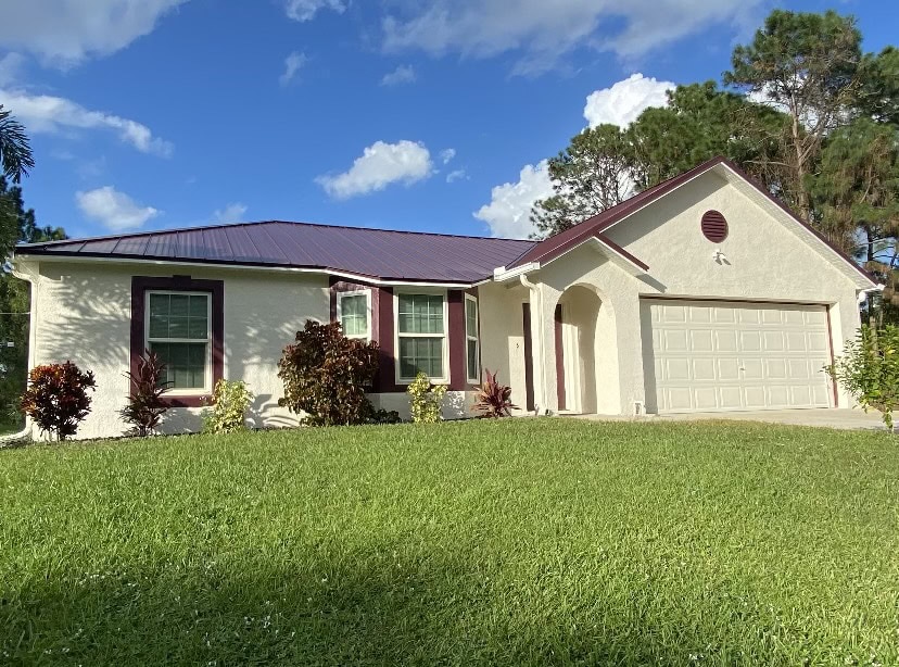 Single-story house with a maroon metal roof, cream-colored stucco walls, maroon trim, and a two-car garage—expertly finished by Stucco Services Central Florida. Surrounded by green grass and shrubs under a blue sky with scattered clouds.