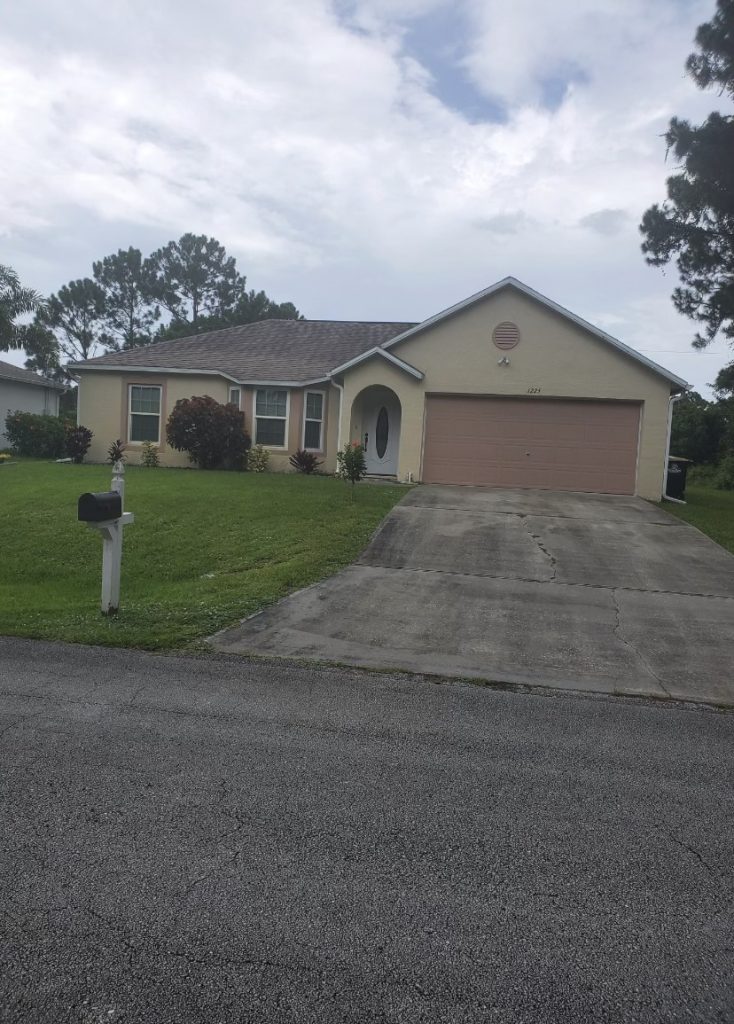 Single-story beige house with a two-car garage, sloped driveway, green lawn, and black mailbox at the edge of the yard. Stucco Services Central Florida enhances its curb appeal. Trees and a cloudy sky are visible in the background.