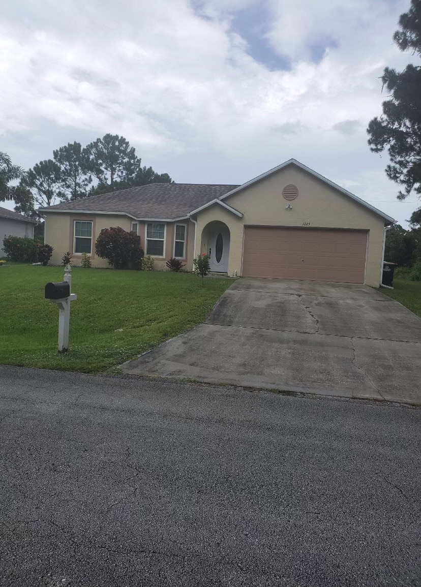 Single-story beige house with a two-car garage, sloped driveway, green lawn, and black mailbox at the edge of the yard. Stucco Services Central Florida enhances its curb appeal. Trees and a cloudy sky are visible in the background.
