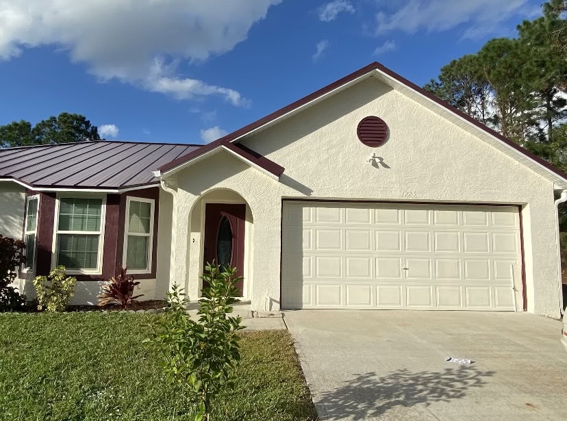 A single-story beige house with a maroon roof and trim features quality stucco services Central Florida trusts, a two-car garage, and a small front yard with grass and shrubs, all under a blue sky with scattered clouds.