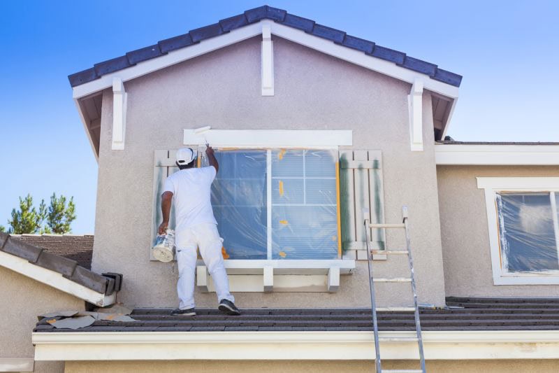 A painter in white clothing stands on the roof, painting the exterior window trim of a beige house. A ladder leans against the home, which features expert stucco services Central Florida residents trust; windows are covered with protective plastic.