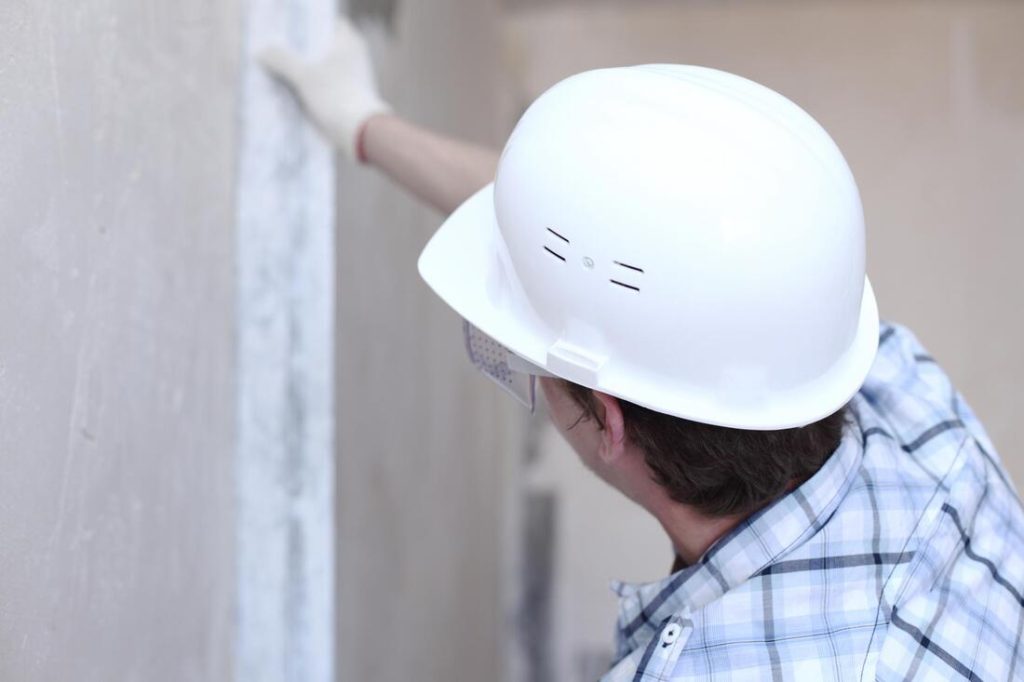 A person wearing a white hard hat, safety glasses, and a plaid shirt is working on a wall, using a tool to smooth or inspect the surface—an example of quality workmanship from Stucco Services Central Florida. The person’s face is not visible.