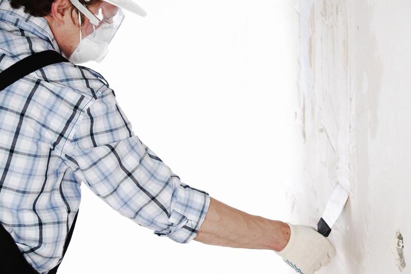A person wearing a hard hat, safety goggles, gloves, and a plaid shirt uses a putty knife to spread plaster or filler on a white wall, demonstrating the expert craftsmanship of Stucco Services Central Florida.