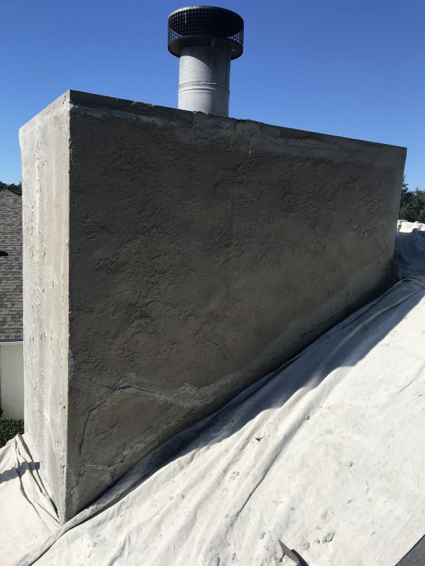 A large, unevenly stuccoed chimney with a metal cap sits atop a sloped roof covered by a protective tarp, under a clear blue sky—a testament to the expert touch of Stucco Services Central Florida.