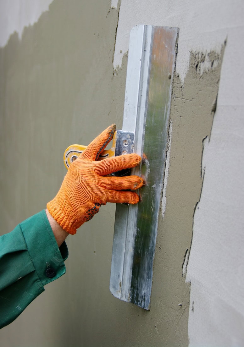 A person wearing an orange glove uses a metal trowel to smooth fresh cement or plaster on a wall, showcasing quality craftsmanship for Stucco Services Central Florida.