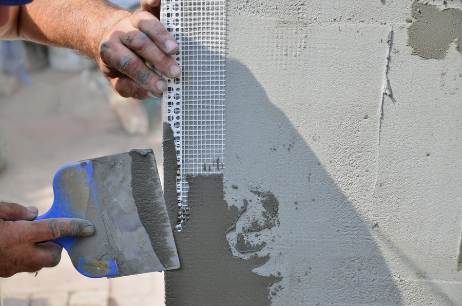 A person applies gray plaster to a wall with a trowel, covering a section of white mesh reinforcement at the corner, showcasing expert Stucco Services Central Florida.