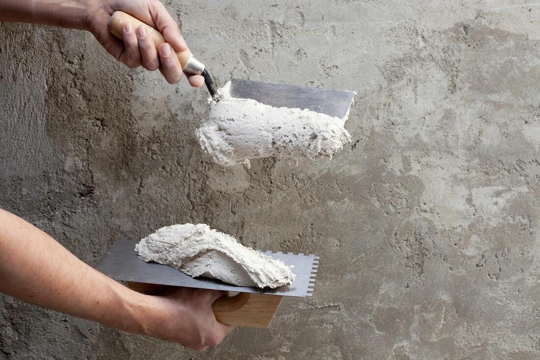 Close-up of hands holding two trowels while applying plaster onto a rough concrete wall, showcasing expert technique from Stucco Services Central Florida as a mound of plaster is transferred from one trowel to the other.
