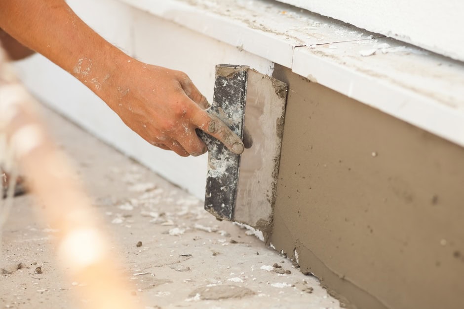 A person’s hand uses a metal trowel to smooth fresh cement or plaster on the exterior wall of a building—typical of Stucco Services Central Florida. Some debris and white tiles are visible nearby.
