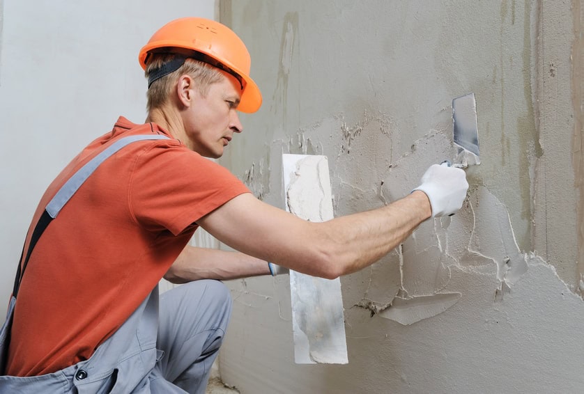 A construction worker wearing an orange hard hat and gloves is applying plaster to a wall with a trowel and a plastering tool, showcasing the quality craftsmanship of Stucco Services Central Florida.