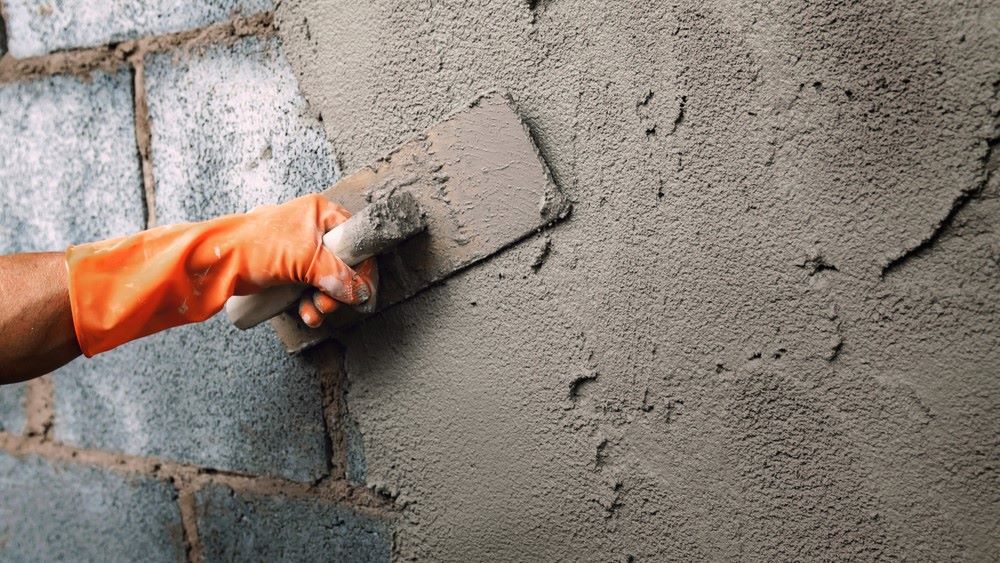A person wearing an orange glove uses a trowel to apply and smooth fresh cement plaster on a rough brick wall, showcasing expert stucco services in Central Florida.