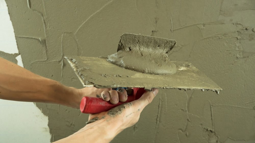A person spreads wet cement onto a wall using a trowel with a red handle, demonstrating a wall-finishing technique often seen in Stucco Services Central Florida.