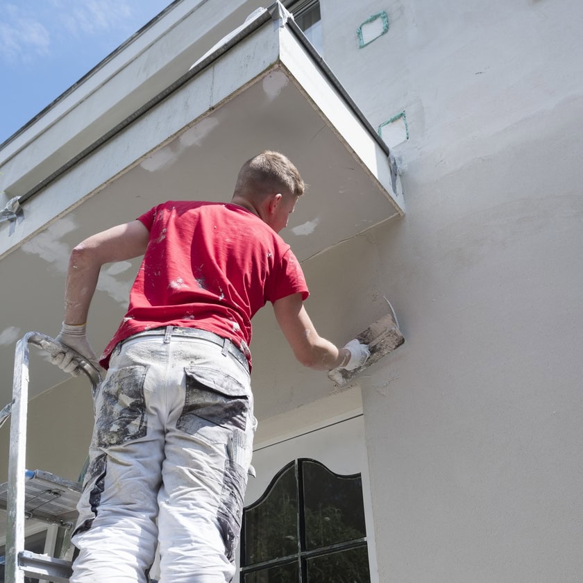 A man standing on a ladder, wearing a red shirt and paint-stained pants, applies plaster or paint to the exterior wall of a building under a balcony on a sunny day—showcasing Stucco Services Central Florida in action.