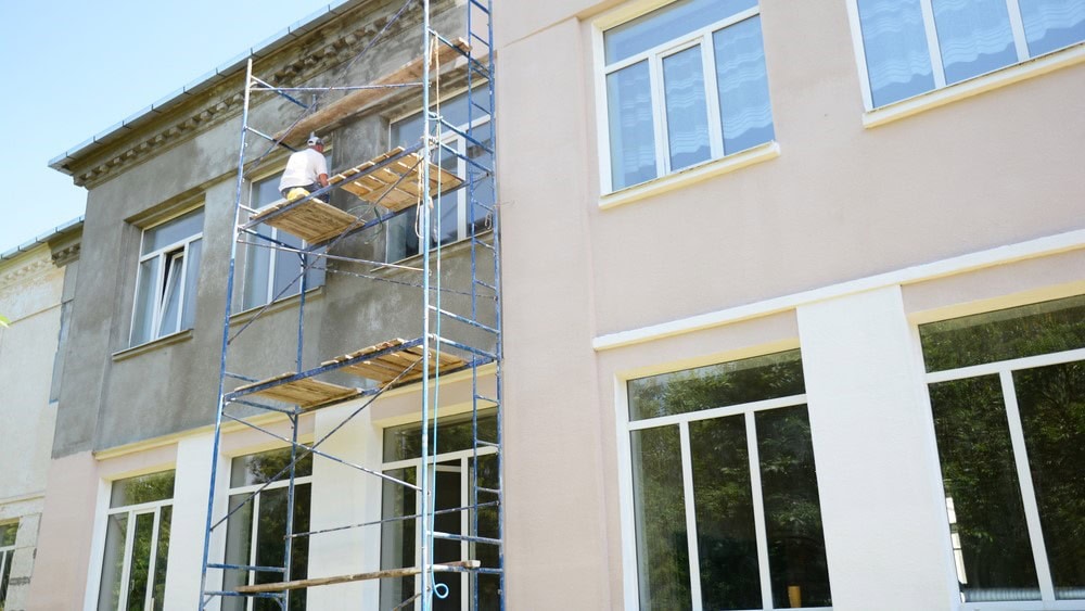 A person stands on scaffolding, painting or repairing the faded exterior wall of a multi-story building. One side, recently updated by Stucco Services Central Florida, is freshly painted a light color, while the other remains weathered.