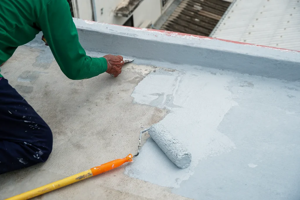 A person applies waterproof coating to a flat concrete roof using a roller and trowel, wearing a green shirt and working near the edge—typical attention to detail seen with Stucco Services Central Florida.