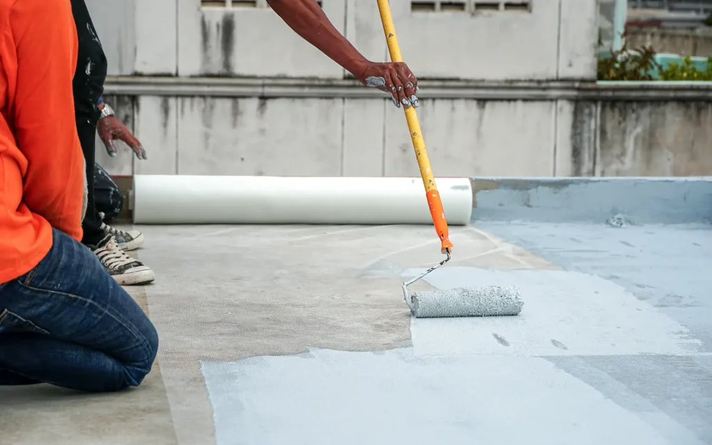 Two people apply a waterproofing membrane to a flat concrete surface using a long-handled paint roller. One person kneels while the other works, showcasing techniques often seen in Stucco Services Central Florida. A white roll sits in the background.