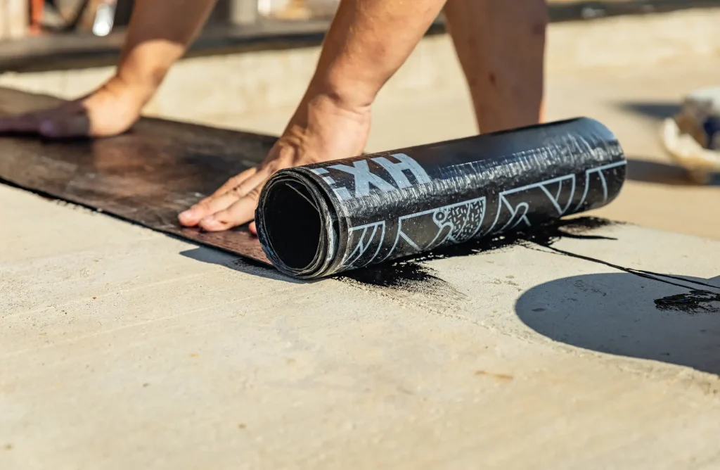 A person’s hands are unrolling and pressing down a roll of black waterproofing membrane onto a flat concrete surface, preparing for roofing or stucco services in Central Florida.